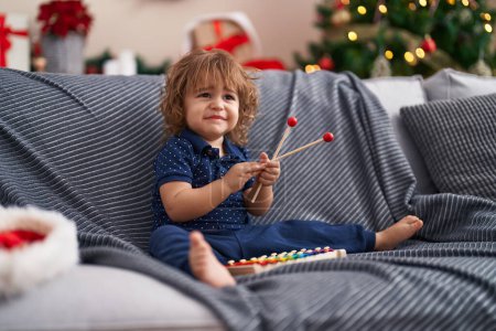 Photo for Adorable hispanic toddler playing xylophone sitting on sofa by christmas gifts at home - Royalty Free Image