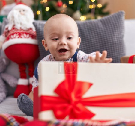Photo for Adorable caucasian baby holding gift sitting on sofa by christmas tree at home - Royalty Free Image