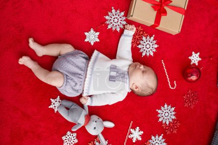 Foto de Adorable bebé caucásico acostado en el suelo con decoración navideña durmiendo sobre un fondo rojo aislado - Imagen libre de derechos