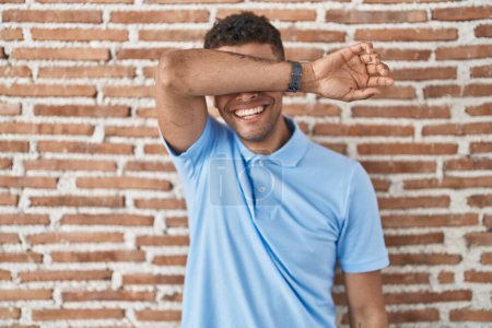 Photo for Brazilian young man standing over brick wall covering eyes with arm smiling cheerful and funny. blind concept. - Royalty Free Image