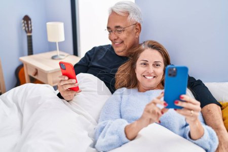 Téléchargez les photos : Couple homme / femme d'âge moyen utilisant un smartphone couché sur le lit dans la chambre - en image libre de droit