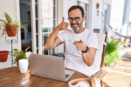 Foto de Hombre de mediana edad utilizando ordenador portátil en casa sonriendo haciendo hablar por teléfono gesto y señalando a usted. Llámame.. - Imagen libre de derechos