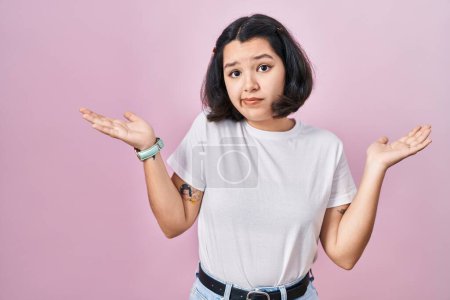 Foto de Young hispanic woman wearing casual white t shirt over pink background clueless and confused expression with arms and hands raised. doubt concept. - Imagen libre de derechos