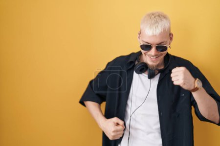 Téléchargez les photos : Jeune homme caucasien portant des lunettes de soleil debout sur fond jaune dansant heureux et joyeux, souriant décontracté et confiant écoutant de la musique - en image libre de droit