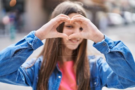 Téléchargez les photos : Adorable fille souriant confiant faire geste de coeur avec les mains à la rue - en image libre de droit