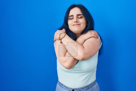 Photo for Young modern girl with blue hair standing over blue background hugging oneself happy and positive, smiling confident. self love and self care - Royalty Free Image