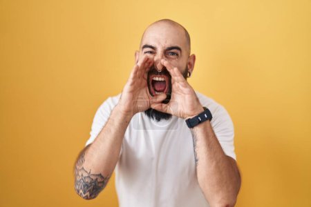 Téléchargez les photos : Young hispanic man with beard and tattoos standing over yellow background shouting angry out loud with hands over mouth - en image libre de droit