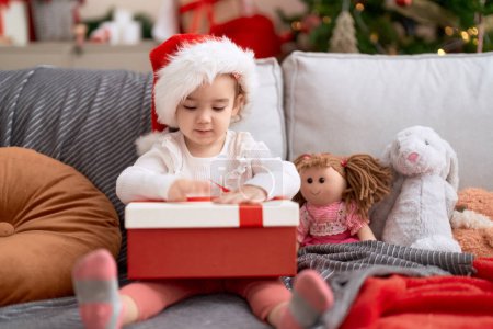 Téléchargez les photos : Adorable fille caucasienne tenant cadeau assis sur le canapé par arbre de Noël à la maison - en image libre de droit