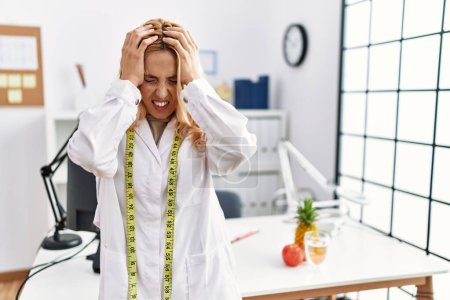 Foto de Beautiful blonde nutritionist woman at dietitian clinic suffering from headache desperate and stressed because pain and migraine. hands on head. - Imagen libre de derechos