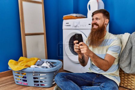 Foto de Joven pelirrojo sonriendo confiado usando smartphone en la lavandería - Imagen libre de derechos