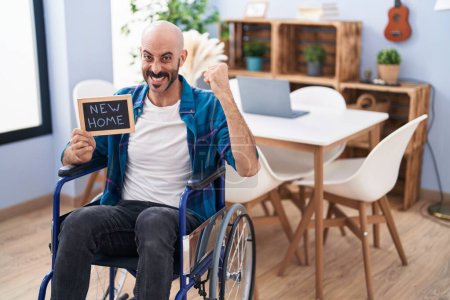 Téléchargez les photos : Homme hispanique avec barbe assis sur fauteuil roulant à la nouvelle maison criant fier, célébrant la victoire et le succès très excité avec le bras levé - en image libre de droit