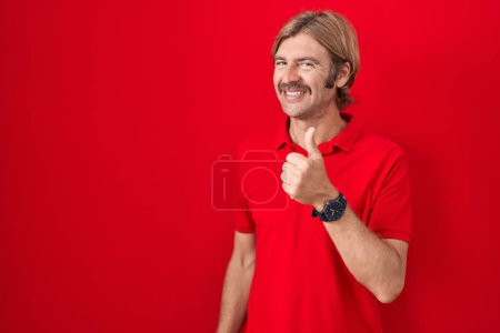 Foto de Caucasian man with mustache standing over red background doing happy thumbs up gesture with hand. approving expression looking at the camera showing success. - Imagen libre de derechos