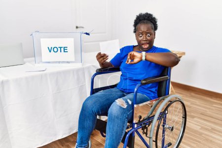 Foto de Mujer africana joven sentada en silla de ruedas votando poniendo sobre en las urnas mirando el tiempo del reloj preocupada, temerosa de llegar tarde - Imagen libre de derechos