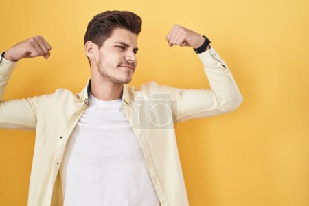 Téléchargez les photos : Jeune homme hispanique debout sur fond jaune montrant les muscles des bras souriant fier. concept de fitness. - en image libre de droit