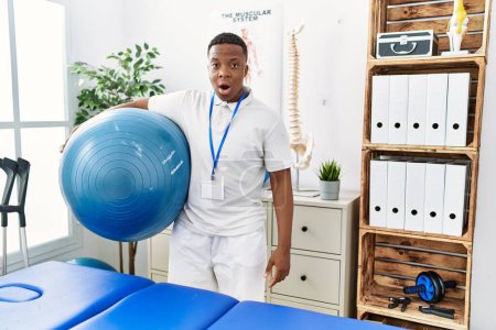 Téléchargez les photos : Jeune homme africain de physiothérapie tenant le ballon pilates à la clinique de réadaptation effrayé et étonné avec la bouche ouverte pour surprise, visage incrédule - en image libre de droit
