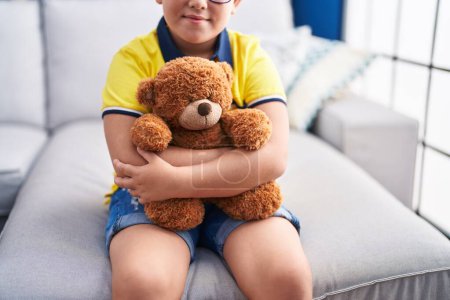 Photo for Adorable hispanic boy hugging teddy bear sitting on sofa at home - Royalty Free Image