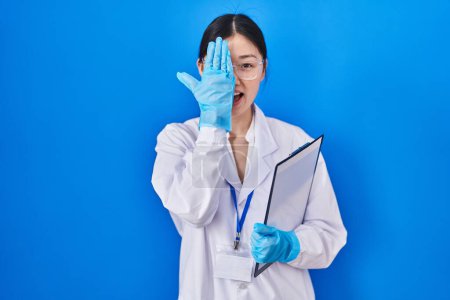 Téléchargez les photos : Jeune femme chinoise travaillant au laboratoire scientifique couvrant un œil avec la main, sourire confiant sur le visage et émotion surprise. - en image libre de droit