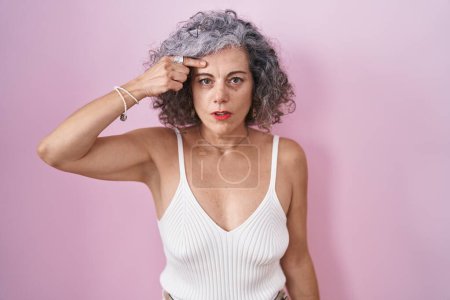 Foto de Mujer de mediana edad con el pelo gris de pie sobre el fondo rosa que apunta infeliz a la espinilla en la frente, fea infección de la espinilla. acné y problemas de piel - Imagen libre de derechos
