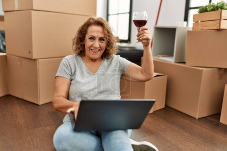 Photo for Middle age caucasian woman using laptop and drinking wine sitting on the floor at new home. - Royalty Free Image
