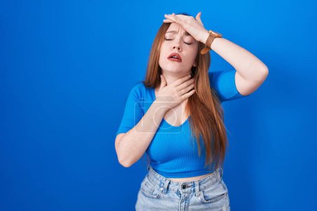 Foto de Redhead woman standing over blue background touching forehead for illness and fever, flu and cold, virus sick - Imagen libre de derechos