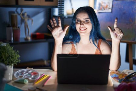 Foto de Joven chica moderna con el pelo azul sentado en el estudio de arte con el ordenador portátil por la noche mostrando y apuntando hacia arriba con los dedos número siete mientras sonríe confiado y feliz. - Imagen libre de derechos