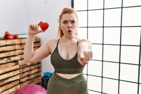 Photo for Young redhead woman holding heart at the gym pointing with finger to the camera and to you, confident gesture looking serious - Royalty Free Image
