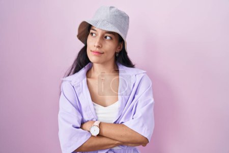Foto de Young hispanic woman standing over pink background wearing hat looking to the side with arms crossed convinced and confident - Imagen libre de derechos
