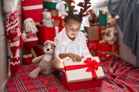 Téléchargez les photos : Adorable hispanique tout-petit déballer cadeau de Noël assis sur le sol à la maison - en image libre de droit
