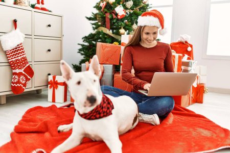 Téléchargez les photos : Jeune femme caucasienne utilisant ordinateur portable assis avec chien par arbre de Noël à la maison - en image libre de droit