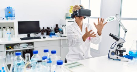 Foto de Mujer rubia de mediana edad vistiendo uniforme científico usando gafas vr trabajando en el laboratorio - Imagen libre de derechos
