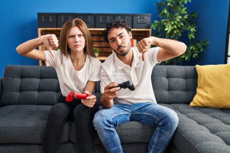 Photo for Young couple playing video game holding controller at home with angry face, negative sign showing dislike with thumbs down, rejection concept - Royalty Free Image