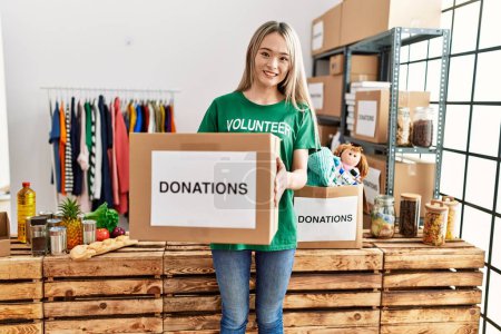 Photo for Young chinese woman wearing volunteer uniform holding donations box at charity center - Royalty Free Image