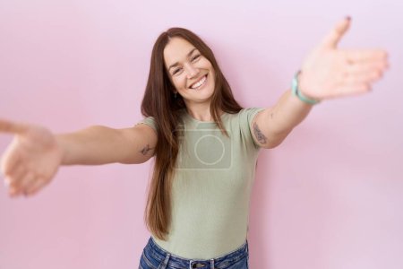 Téléchargez les photos : Belle femme brune debout sur fond rose regardant la caméra souriant à bras ouverts pour se faire câliner. expression joyeuse embrassant le bonheur. - en image libre de droit
