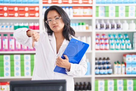 Photo for Asian young woman working at pharmacy drugstore holding clipboard with angry face, negative sign showing dislike with thumbs down, rejection concept - Royalty Free Image