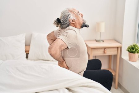 Foto de Middle age grey-haired man suffering for back injury sitting on bed at bedroom - Imagen libre de derechos
