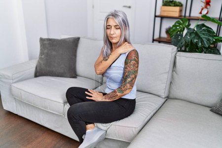 Téléchargez les photos : Femme aux cheveux gris d'âge moyen avec douleur au cou assise sur le canapé à la maison - en image libre de droit