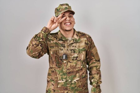 Foto de Joven árabe vestido con camuflaje uniforme del ejército haciendo símbolo de paz con los dedos sobre la cara, sonriendo alegre mostrando la victoria - Imagen libre de derechos