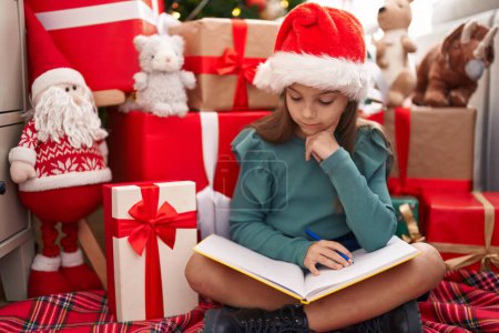 Foto de Adorable chica hispana leyendo libro sentado en el suelo por árbol de Navidad en casa - Imagen libre de derechos