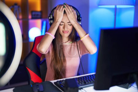 Foto de Mujer hispana joven jugando videojuegos con dolor de cabeza desesperada y estresada por el dolor y la migraña. manos en la cabeza. - Imagen libre de derechos