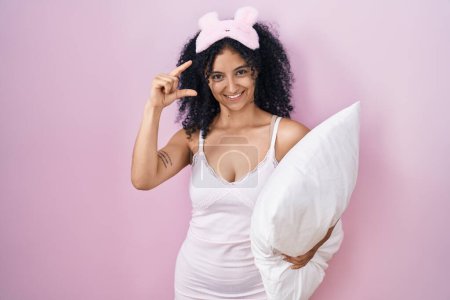 Téléchargez les photos : Hispanic woman with curly hair wearing sleep mask and pajama holding pillow smiling and confident gesturing with hand doing small size sign with fingers looking and the camera. measure concept. - en image libre de droit