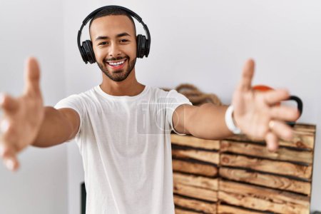 Téléchargez les photos : Homme afro-américain écoutant de la musique à l'aide d'écouteurs à la salle de gym en regardant la caméra souriant à bras ouverts pour étreindre. expression joyeuse embrassant le bonheur. - en image libre de droit
