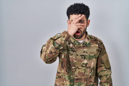 Foto de Hombre árabe vistiendo camuflaje uniforme del ejército asomándose en shock cubriendo cara y ojos con la mano, mirando a través de los dedos con expresión avergonzada. - Imagen libre de derechos