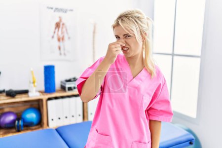 Photo for Young caucasian woman working at pain recovery clinic smelling something stinky and disgusting, intolerable smell, holding breath with fingers on nose. bad smell - Royalty Free Image