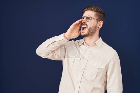 Foto de Hispanic man with beard standing over blue background shouting and screaming loud to side with hand on mouth. communication concept. - Imagen libre de derechos
