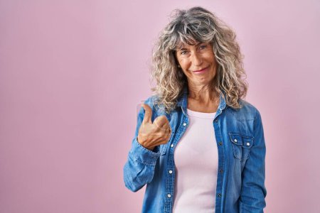 Photo for Middle age woman standing over pink background doing happy thumbs up gesture with hand. approving expression looking at the camera showing success. - Royalty Free Image