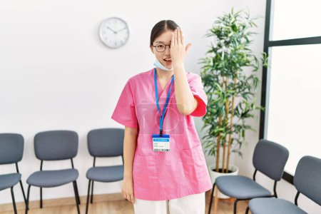 Téléchargez les photos : Jeune infirmière asiatique femme à la salle d'attente médicale couvrant un oeil avec la main, sourire confiant sur le visage et l'émotion surprise. - en image libre de droit