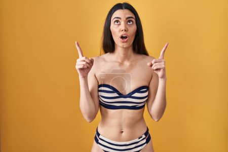 Téléchargez les photos : Jeune femme brune portant un bikini sur fond jaune étonné et surpris regardant vers le haut et pointant avec les doigts et les bras levés. - en image libre de droit