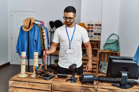 Téléchargez les photos : Jeune homme hispanique commerçant souriant confiant nettoyage poussière au magasin de vêtements - en image libre de droit