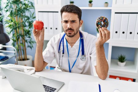 Foto de Young hispanic dietitian man holding doughnut and apple skeptic and nervous, frowning upset because of problem. negative person. - Imagen libre de derechos