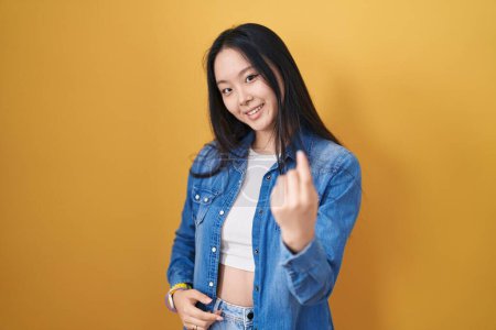 Young asian woman standing over yellow background beckoning come here gesture with hand inviting welcoming happy and smiling 